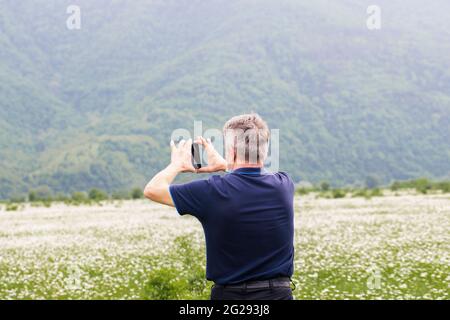 Kaukasischer Mann nimmt ein weißes Feld von Wildblumen-Gänseblümchen auf das Telefon. Kamillen auf dem Hintergrund von Bergen. Stockfoto