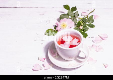 Tee aus Hagebutten Blüten. Sommer Vitamingetränk, hartes Licht, dunkler Schatten. Weißer Holzhintergrund, Kopierraum Stockfoto