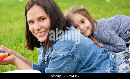 Mama lächelt mit einer kleinen Tochter 4 Jahre auf dem Gras liegen, Ruhe im Sommer, Blick auf die Kamera. Mutter hält ein Spielzeug Pop IT, ein glückliches Mädchen, das in einer Mutter auf dem Rücken liegt Stockfoto