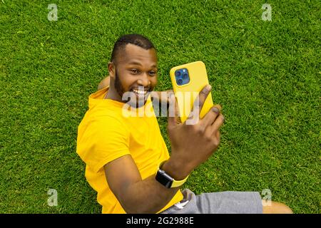 Schöner schwarzer afro amerikanischer junger Mann mit Mobiltelefon in der Hand im Park oder im Stadion auf grünem Gras sitzend, Online-Geschäftstreffen oder Stockfoto