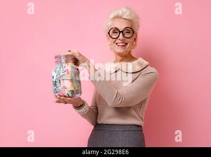 Lächelnde Großmutter zeigt ihr, wie sie im Glas Geld spart. Foto von gutherzigen älteren Frau trägt Brillen auf rosa Hintergrund. Stockfoto