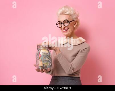 Lächelnde Großmutter zeigt ihr, wie sie im Glas Geld spart. Foto von gutherzigen älteren Frau trägt Brillen auf rosa Hintergrund. Stockfoto