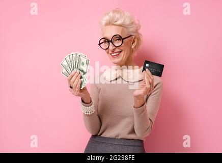 Lächelnde Großmutter zeigt Bargeld und Kreditkarte. Foto von gutherzigen älteren Frau auf rosa Hintergrund. Stockfoto