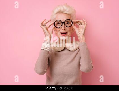 Überfröhliche Großmutter lacht über etwas Lustiges. Foto einer freundlichen älteren Frau in einer Brille, die auf einem rosa Hintergrund die Kamera anschaut. Stockfoto