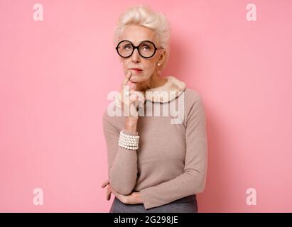 Nachdenkliche Oma, die tief in Gedanken ist, kurvt die Lippen, hält das Kinn fest und schaut weg. Foto von gutherzigen älteren Frau trägt Brillen auf rosa Hintergrund. Stockfoto