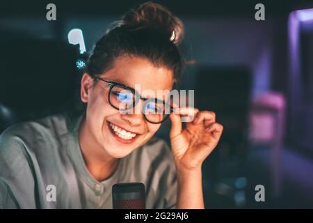 Eine schöne Frau in einer Brille, die als Radiomoderatorin arbeitet und hinter dem Mikrofon lächelt. Sie nimmt ein Interview und hört einem anderen Redner mit Freude zu Stockfoto