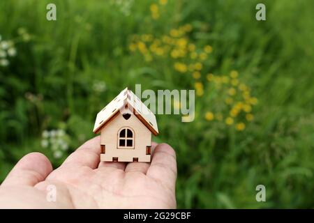 Holzhaus in männlicher Hand auf dem Hintergrund der Sommerwiese mit wilden Blumen. Immobilienmakler, Kauf oder Vermietung Haus in ökologisch sauberen Bereich Stockfoto