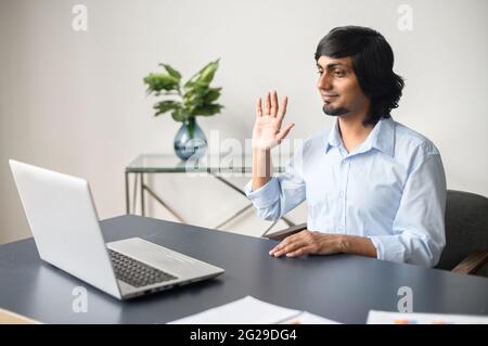 Lächelnder indischer, männlicher Büroangestellter, der online über Video Connect auf dem Laptop spricht, nimmt an einem virtuellen Meeting Teil und winkt in die Webcam Stockfoto
