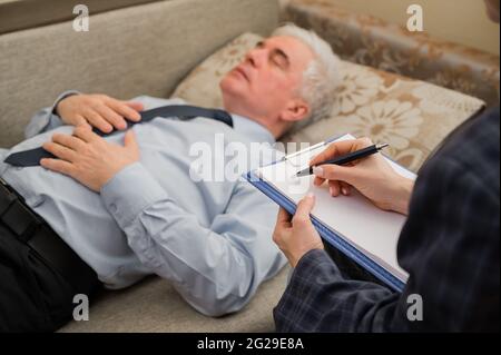 Ein älterer Mann liegt während eines Termins mit einem Psychotherapeuten auf einer Couch. Ein älterer männlicher Patient in einer individuellen Konsultation mit einer Frau Stockfoto