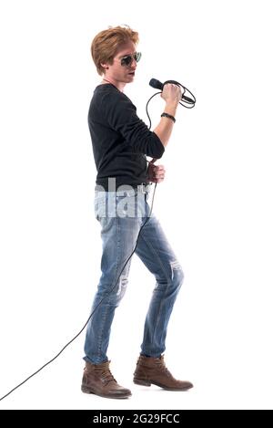 Seitenansicht eines hübschen jungen Rockers, der das Mikrofon singt und hält. Ganzkörperlänge isoliert auf weißem Hintergrund. Stockfoto