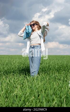 Das kleine Mädchen mit Sonnenbrille richtet ihr Haar auf einem grünen Grasfeld vor dem Hintergrund eines bewölkten Himmels. Stockfoto