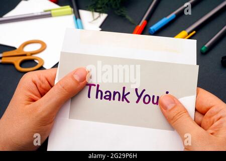 Frau Hände nehmen aus grauem Papier mit Worten Danke aus dem Umschlag. Büromaterial im Hintergrund. Stockfoto