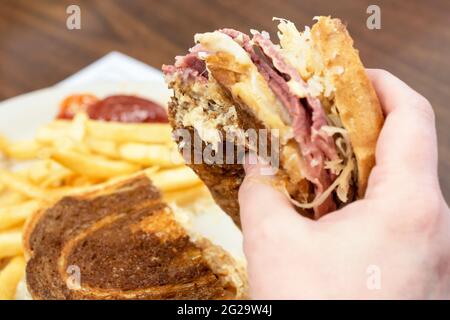 Ein Patron hält ein Rueben-Sandwich in der einen Hand, während die andere Hälfte des Sandwiches und pommes frites im Hintergrund verschwommen sind. Stockfoto