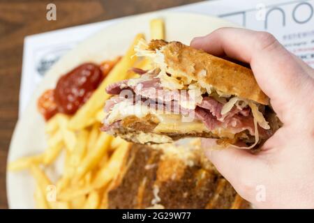 Ein Patron hält ein Rueben-Sandwich in der einen Hand, während die andere Hälfte des Sandwiches und pommes frites im Hintergrund verschwommen sind. Stockfoto