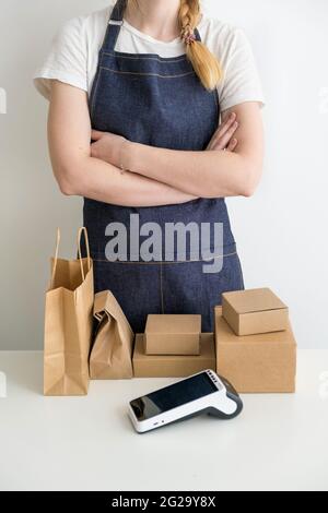 Junge Frau trägt Schürze mit den Armen gekreuzt mit Kartons, Pappverpackungen und modernen Zahlungsterminal am Tisch Stockfoto