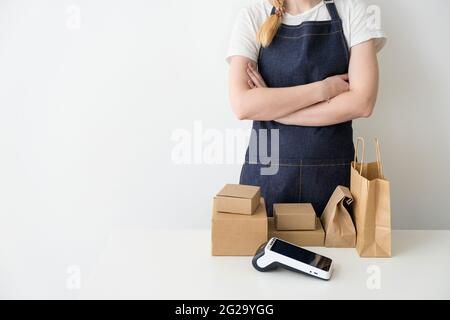 Junge Frau trägt eine Jeanschürze mit gekreuzten Armen, die am Tisch stehen, mit Kartons, Pappverpackungen und einem modernen Zahlungsterminal Stockfoto