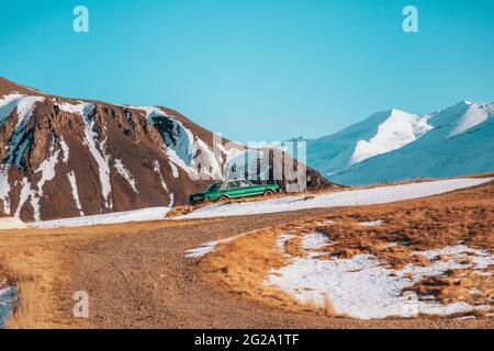 Grünes Auto am Hang eines Hügels zwischen schneebedeckten mächtigen Bergen mit klarem blauen Himmel auf dem Hintergrund in Island Stockfoto