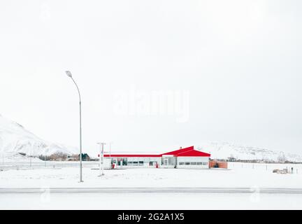 Modernes Haus mit rotem Dach auf leerem Straßenrand in der Nähe von Straßenlaternen inmitten von endlosen verschneiten Bergen mit weißem Himmel auf dem Hintergrund in Island Stockfoto