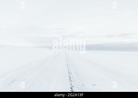 Leere endlose schneebedeckte Straße zwischen schneebedeckten weißen Feldern mit bewölktem Himmel auf dem Hintergrund in Island Stockfoto