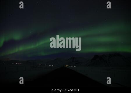 Gerade Straße durch verschneite Landschaft in Richtung Berge gegen Himmel mit grünen Polarlichtern in der Winternacht in Island Stockfoto