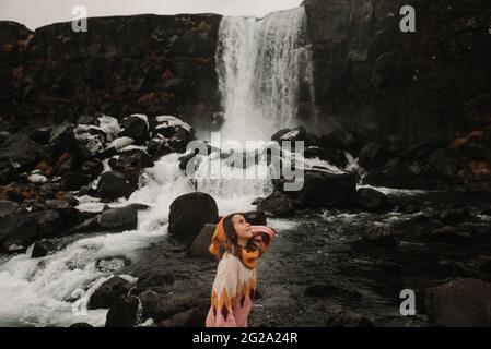 Junges Mädchen mit Blick weg in stilvollem Outfit neben majestätischen Blick auf wunderbare Wasserfall in isländischen Landschaft Stockfoto