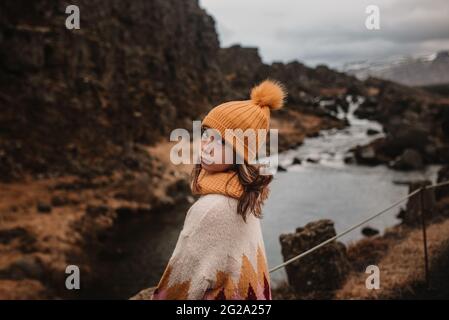 Rückansicht eines jungen Mädchens in warmen Kleidern, das auf einer Brücke steht und an einem langweiligen Tag in der isländischen Landschaft die Kamera neben dem ruhigen Fluss anschaut Stockfoto