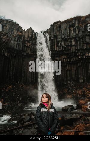 Junges Mädchen mit Blick weg in stilvollem Outfit neben majestätischen Blick auf wunderbare Wasserfall in isländischen Landschaft Stockfoto