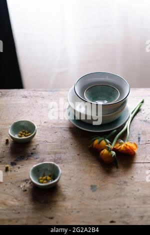 Hoher Winkel von frischen, leuchtend orangefarbenen Ranunculus-Blumen und Kapern in kleinen Keramikschüsseln auf braunem Holztisch im hellen Raum Stockfoto