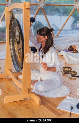 Seitenansicht einer ausgeglichenen Frau in weißen Kleidern, die Gong trifft, während regungslose Menschen in Shavasana mit geschlossenen Augen liegen und mit einer Decke über Yoga bedeckt sind Stockfoto