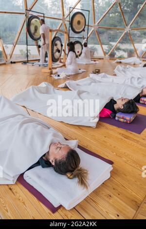 Seitenansicht einer ausgeglichenen Frau in weißen Kleidern, die Gong trifft, während regungslose Menschen in Shavasana mit geschlossenen Augen liegen und mit einer Decke über Yoga bedeckt sind Stockfoto