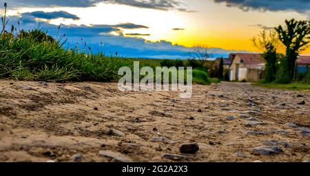 Panoramaaufnahme eines trockenen schmalen Pfades durch grasbewachsene Felder in der Nähe von Bauernhäusern unter einem Sonnenuntergangshimmel Stockfoto