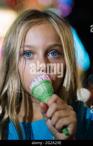 Kleines blondes Mädchen, das nachts im Vergnügungspark ein leckeres Eis isst Stockfoto