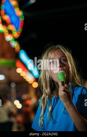 Kleines blondes Mädchen, das nachts im Vergnügungspark ein leckeres Eis isst Stockfoto