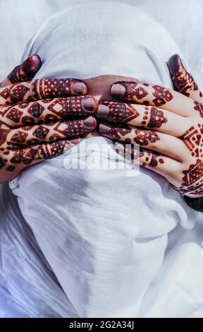 Tätowierte Hände einer nicht erkennbaren Frau, die die Augen einer anonymen arabischen Person mit einem traditionellen weißen Kopftuch bedeckt Stockfoto
