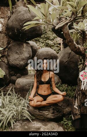 Junge attraktive ethnische Frau im Bikini, die Yoga in einem wunderschönen tropischen Garten praktiziert Stockfoto