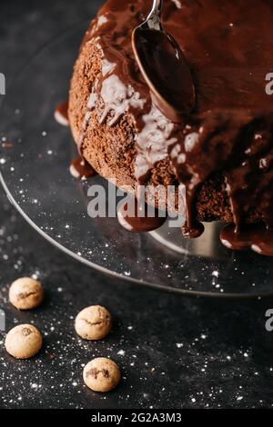 Crop anonyme Person Hand mit Löffel Verbreitung Schokolade Ganache auf hausgemachten Kuchen auf runden Glasständer auf dem Tisch vor schwarzem Hintergrund platziert Stockfoto