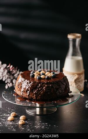 Appetitlich hausgemachter Kuchen mit Schokolade Ganache mit Nüssen und Heidelbeeren auf dem Tisch mit einer Flasche Milch auf schwarzem Hintergrund dekoriert Stockfoto