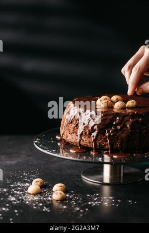 Crop anonyme Hausfrau Anordnung Nüsse auf hausgemachten Kuchen mit Schokolade Belag auf dem Tisch vor schwarzem Hintergrund platziert Stockfoto