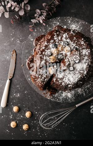 Draufsicht auf leckeren Schokoladenkuchen mit geschnittenem Stück mit Schokoladensirup und Zuckerpulver auf dem Tisch mit Messer und Schneebesen angeordnet Stockfoto
