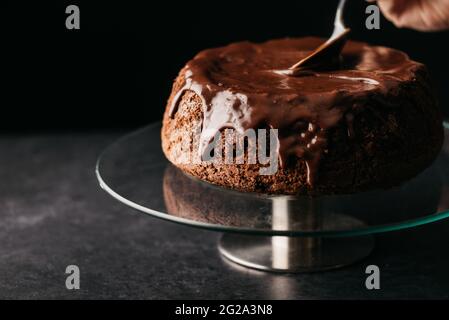 Crop anonyme Person Hand mit Löffel Verbreitung Schokolade Ganache auf hausgemachten Kuchen auf runden Glasständer auf dem Tisch vor schwarzem Hintergrund platziert Stockfoto