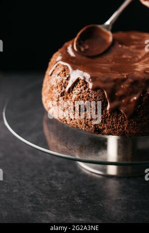 Crop anonyme Person Hand mit Löffel Verbreitung Schokolade Ganache auf hausgemachten Kuchen auf runden Glasständer auf dem Tisch vor schwarzem Hintergrund platziert Stockfoto