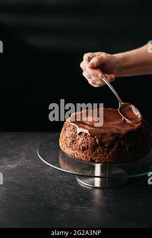 Crop anonyme Person Hand mit Löffel Verbreitung Schokolade Ganache auf hausgemachten Kuchen auf runden Glasständer auf dem Tisch vor schwarzem Hintergrund platziert Stockfoto