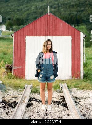 Schöne Frau in trendigen Outfit Blick auf die Kamera, während in der Nähe von roten Scheune in der Landschaft stehen Stockfoto