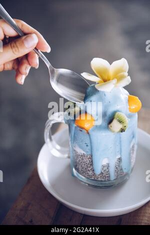 Crop Hand der Frau hält Löffel in der Nähe köstliche frische Dessert mit Früchten in Glas Becher auf Untertasse Stockfoto