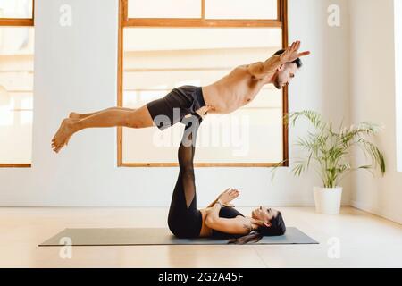 Die junge Frau, die auf einer Yogamatte liegt, hält einen muskulösen Mann auf dem Bein, während der Unterrichtsstunde im Trainingsraum Stockfoto