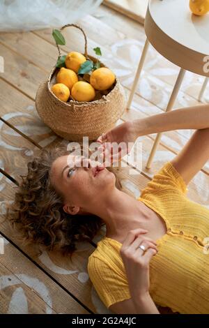 Von oben eine entspannte schlanke Frau, die auf dem Boden neben dem Tisch mit Zitrone im Korb in einem hellen, einfachen Raum liegt und wegschaut Stockfoto