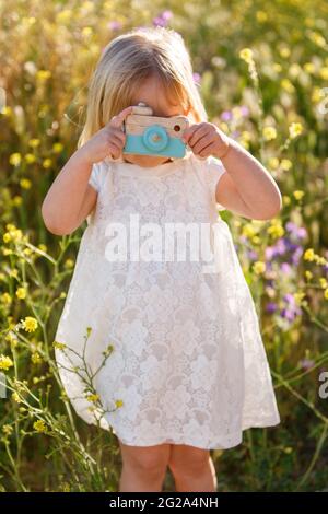 Anonymes kleines Mädchen in weißem Kleid, das die Spielzeugkamera in der Nähe des Gesichts hält, während es an sonnigen Tagen inmitten wilder Blumen auf dem Feld steht Stockfoto