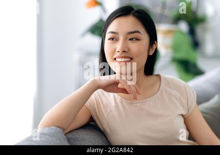 Porträt eines hübschen, gesunden, positiven asiatischen Brünetten-Mädchens, im lässigen T-Shirt, zu Hause auf einem Sofa sitzend, entspannend, wegblickend, vom Urlaub träumend, glücklich lächelnd Stockfoto