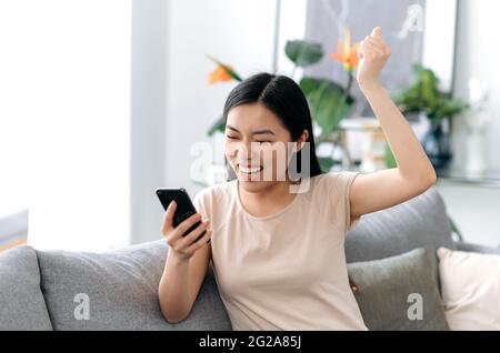 Überglücklich sitzt eine junge Asiatin, modern gekleidet, auf dem Sofa im Wohnzimmer, hält ein Smartphone in der Hand, freut sich über den Sieg oder eine gute Nachricht, lächelt fröhlich und schaut aufs Telefon Stockfoto