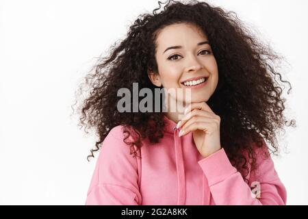 Schönheit und Lifestyle. Junge Frau mit natürlich glühender Haut, lockiges dunkles Haar, lächelnden weißen Zähnen und glücklich, berühren Kinn nachdenklich, haben Stockfoto